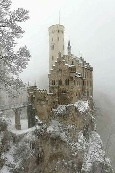 an old castle on top of a mountain covered in snow and surrounded by trees, with a bridge leading to it