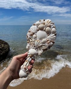 a hand holding up some shells on the beach near the water's edge with waves coming in