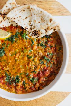 a white bowl filled with lots of food on top of a wooden table next to pita bread