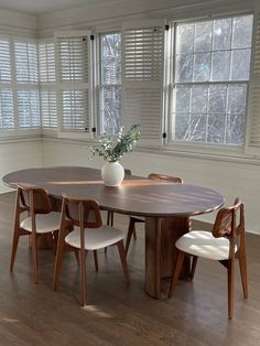 a dining room table with chairs and a vase on it in front of two windows