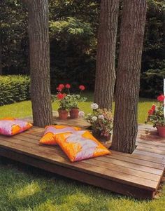 a wooden deck with pillows on it in the middle of some trees and flowers outside