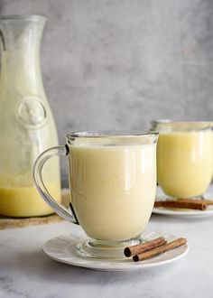 two mugs filled with coffee sit on a plate next to cinnamon sticks and a pitcher