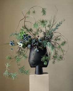 a black vase filled with green and blue flowers on top of a wooden block in front of a wall