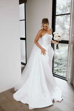 a woman in a white wedding dress standing by a window with her hands on her hips