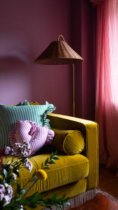 a yellow couch with pillows on it in front of a lamp and striped curtained window