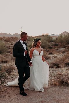 a bride and groom are walking through the desert in their wedding attire, smiling at each other