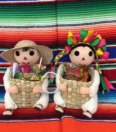 two dolls sitting on top of a colorful blanket holding baskets filled with food and drinks