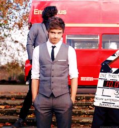 a man standing in front of a red double decker bus holding a sign that says one direction