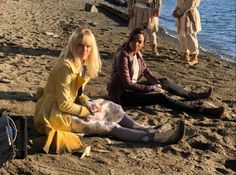 two women sitting on the beach with their feet in the sand while others stand and sit nearby