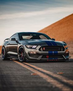 a gray mustang with blue stripes driving down the road in front of a hill side