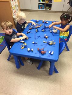 three children sitting at a blue table playing with toy cars and trucks on the floor