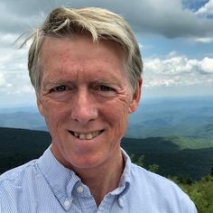 a man with blonde hair and blue shirt smiling at the camera on top of a mountain