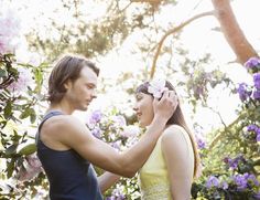 a man and woman standing next to each other in front of trees with purple flowers