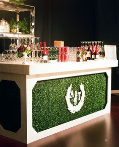 an outdoor bar is decorated with artificial grass and wine glasses on the counter, surrounded by greenery