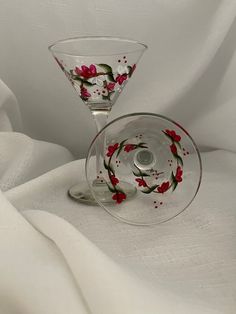 two wine glasses sitting on top of a white cloth covered tablecloth with red flowers painted on them