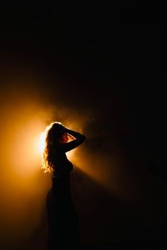 a woman standing in the dark with her hands on her head and light shining behind her