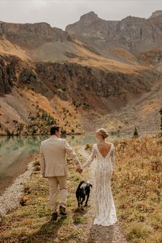 a bride and groom walking with their dog on the side of a mountain trail near a lake