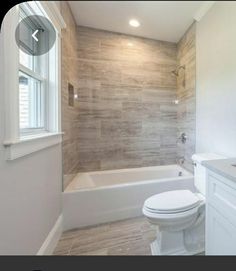 a white toilet sitting next to a bath tub under a window in a bathroom with wood flooring