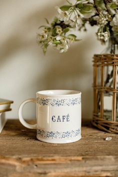 a white coffee mug sitting on top of a wooden table next to a vase with flowers