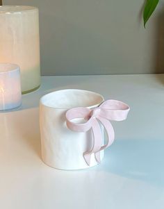 a white candle holder with a pink ribbon tied around it next to a lit candle