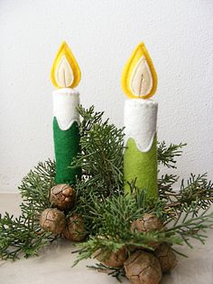 two candles are sitting on top of pine cones and fir needles, decorated with felt