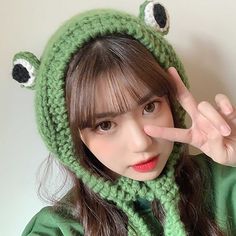 a woman wearing a green frog hat and scarf with eyes drawn on her nose while making a hand sign