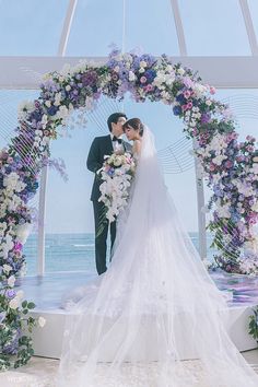 a bride and groom standing in front of an arch decorated with purple, white and blue flowers