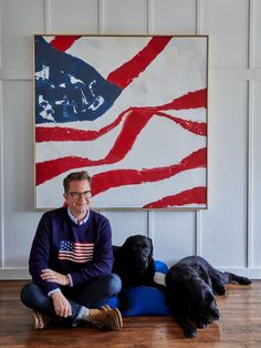 a man sitting on the floor next to two dogs with an american flag painting behind him