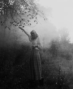 a woman is standing in the woods with her arms out and reaching for leaves from a tree