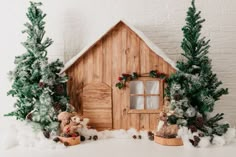 a small wooden house surrounded by snow and christmas trees, with a teddy bear in front