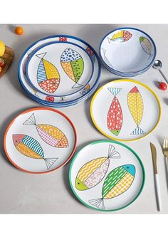 four colorful plates with fish designs on them next to utensils and fruit bowl