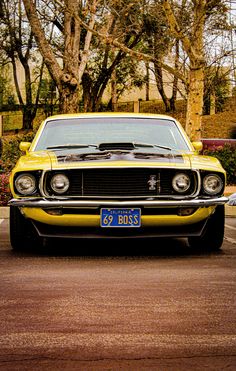 a yellow muscle car parked in a parking lot next to some trees and bushes on a sunny day