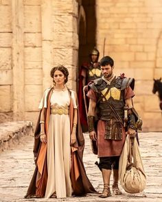 a man and woman dressed in roman costumes walking down a cobblestone street with horses behind them