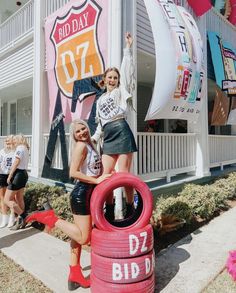 two girls are posing in front of a building with giant tires on the sidewalk and banners behind them