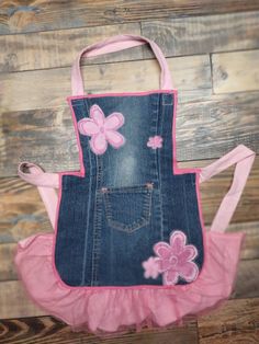 a denim apron with pink flowers on the front and bottom, hanging from a wooden wall