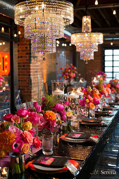 a long table is set with pink and orange flowers in vases, silverware, and crystal chandeliers