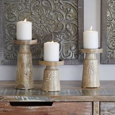 three white candles sitting on top of a wooden table next to a wall mounted painting