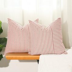 two red and white striped pillows on a wooden shelf next to a plant in front of a window