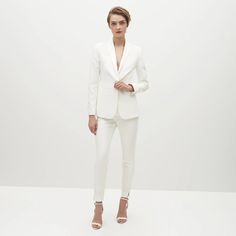 a woman in a white suit and heels standing on a white surface with her hands behind her back