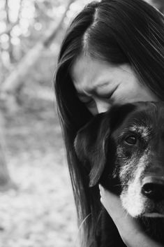 a black and white photo of a woman hugging her dog's face with trees in the background