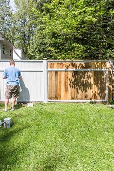 a man standing next to a white fence in the grass near a house and trees