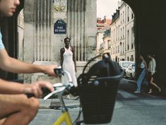 two people riding bikes on a city street with buildings in the background and one person wearing a white dress