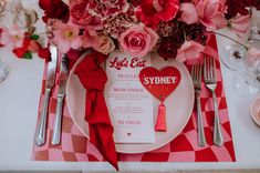 A table setting with red and pink checkerboard placemat, a pink plate with a red napkin, menu and red heart shaped name card with a red tassel with red and pink flowers at the top of the image Wedding Poker Table, Pink Red Table Setting, Valentine Wedding Theme, Valentines Themed Wedding, Pink And Red Table Setting, Valentines Elopement