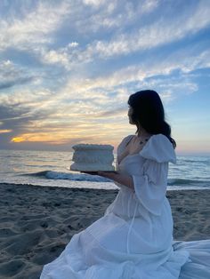 a woman in a white dress holding a cake on the beach at sunset or dawn