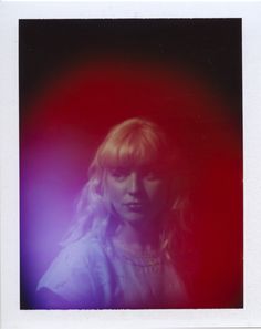 a woman with blonde hair standing in front of a red and blue background, looking at the camera