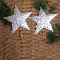 two silver star ornaments hanging on a brown surface next to pine needles and red beads