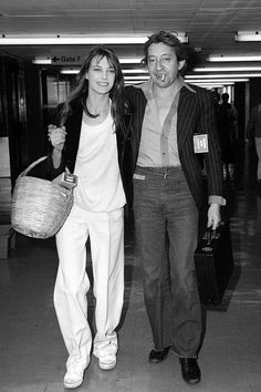 a man and woman walking through an airport with luggage in their hands, smiling at the camera