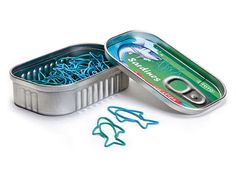 an open tin with fish hooks in it on a white surface next to some paper clips