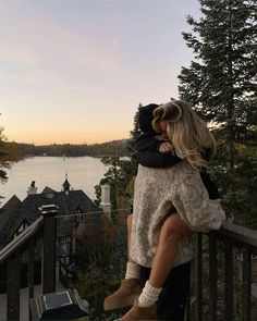 two people hugging each other while standing on a balcony overlooking a lake and forest at sunset