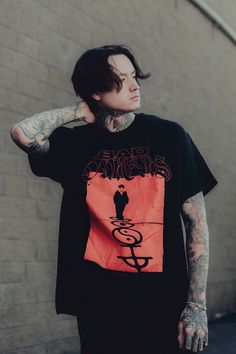 a man with tattoos standing in front of a brick wall wearing a black t - shirt